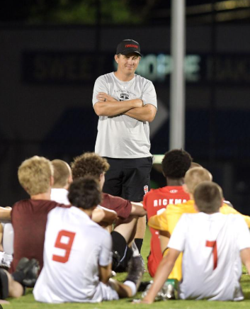 A man in a cap stands with his arms crossed in front of seated Wesleyan soccer players.